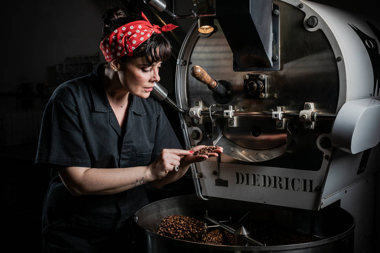 A imagem mostra uma mulher com um lenço vermelho de bolinhas brancas, usando um macacão preto, examinando grãos de café em sua mão enquanto está próxima a uma torrefadora de café da marca Diedrich. A torrefadora é de metal prateado e possui um design industrial. O ambiente é escuro, com foco na mulher e na máquina.