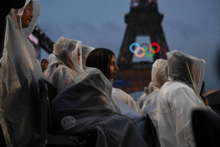A imagem mostra um grupo de pessoas sentadas, usando capas de chuva brancas, em um ambiente ao ar livre. Ao fundo, a Torre Eiffel é visível, iluminada, com os anéis olímpicos projetados. O céu está nublado, sugerindo um clima chuvoso.