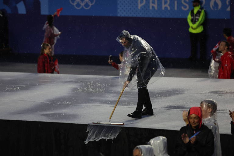 A imagem mostra uma pessoa vestindo uma capa de chuva, utilizando um rodo para limpar uma superfície molhada durante um evento olímpico. Ao fundo, há outras pessoas vestindo roupas coloridas e um segurança em uniforme. O ambiente parece ser um espaço preparado para uma apresentação ou cerimônia.