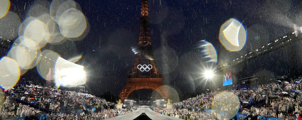 A imagem mostra uma cerimônia olímpica à noite, com a Torre Eiffel ao fundo. O ambiente está iluminado por luzes brilhantes e há uma grande multidão de espectadores. A cena é embaçada por gotas de água, sugerindo que pode estar chovendo. A estrutura de um palco ou rampa é visível em primeiro plano, com a Torre Eiffel destacando-se no centro.

