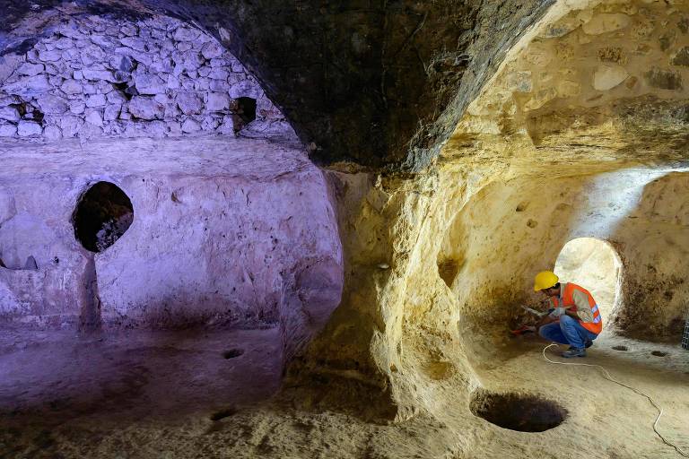 A imagem mostra o interior de uma caverna com formações rochosas. À esquerda, há uma parede de pedra clara com um buraco circular. À direita, uma área mais ampla da caverna, onde uma pessoa está agachada, vestindo roupas coloridas. A iluminação na caverna é variada, com tons de roxo e amarelo, destacando as texturas das rochas.
