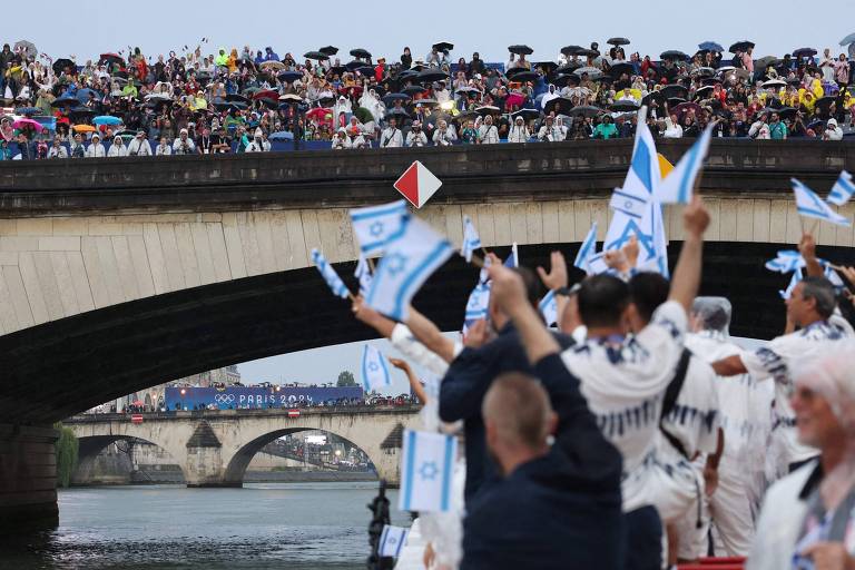 A imagem mostra uma grande multidão reunida em uma ponte sobre um rio, com muitas pessoas segurando bandeiras de Israel e usando guarda-chuvas. Abaixo da ponte, há um grupo de pessoas em um barco, também segurando bandeiras. Ao fundo, é possível ver uma faixa com a inscrição 'PARIS 2024', indicando que o evento está relacionado aos Jogos Olímpicos. O clima parece nublado e chuvoso.
