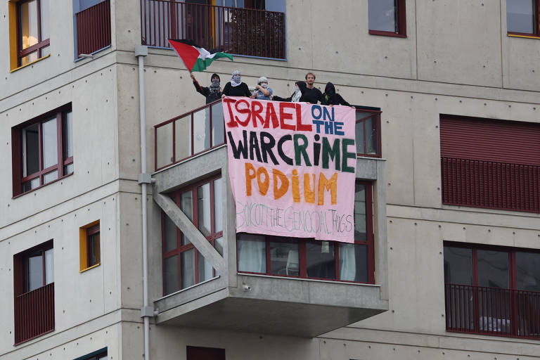 A imagem mostra um grupo de pessoas em uma varanda de um prédio, segurando uma bandeira e um grande banner. O banner tem a inscrição 'ISRAEL ON THE WAR CRIME PODIUM' em letras grandes e coloridas, com a palavra 'WAR CRIME' em laranja. A bandeira que está sendo segurada é de cores verde, vermelho e preto.
