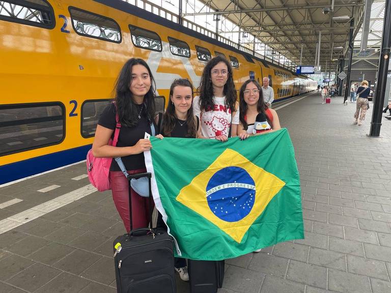 Quatro garotas adolescentes estão em frente a um trem parado na estação. Elas seguram uma bandeira do Brasil aberta