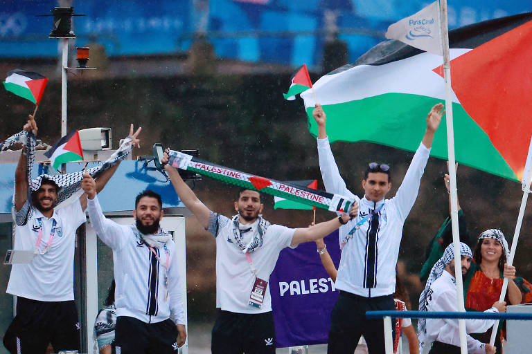 Um grupo de atletas palestinos celebra com bandeiras e um cachecol que diz 'PALESTINE'. Eles estão em um ambiente festivo, com alguns usando roupas esportivas e lenços tradicionais. Ao fundo, há uma bandeira palestina grande e um banner com a palavra 'PALESTINE'.

