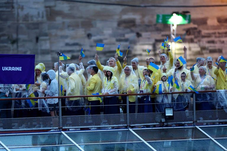 Um grupo de pessoas vestindo capas de chuva transparentes e roupas amarelas está reunido em uma plataforma, segurando bandeiras da Ucrânia. Ao fundo, há uma bandeira roxa com a palavra 'UKRAINE' escrita em letras brancas. O ambiente parece ser um evento ao ar livre, possivelmente sob chuva, com uma estrutura metálica visível na parte inferior da imagem.
