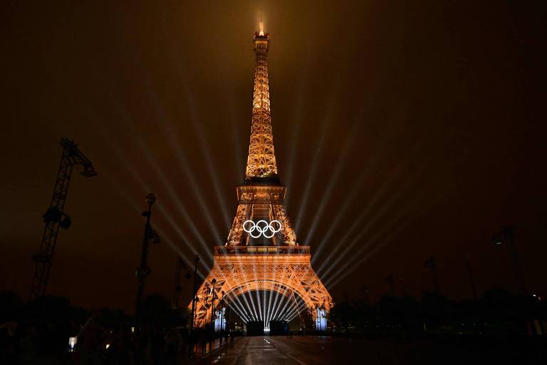 Fotografia mostra a Torre Eiffel no centro, iluminada em tons de amarelo, com o símbolo das Olimpíadas iluminado em branco no centro da estrutura. É noite e todo o resto da imagem está escura