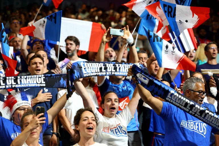 Cenário movimento mostra tum estádio cheio de torcedores. Eles levantam faixas de apoio ao time da França. É possível ver bandeiras da França ao fundo.