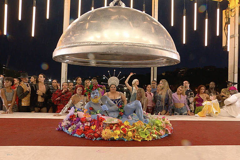 Imagem mostra um evento festivo, com uma grande cúpula prateada ao fundo. No centro, uma pessoa está sentada em um vestido colorido, cercada por flores. Ao redor, um grupo de pessoas aplaude e observa, algumas levantando as mãos. O ambiente é iluminado por luzes penduradas