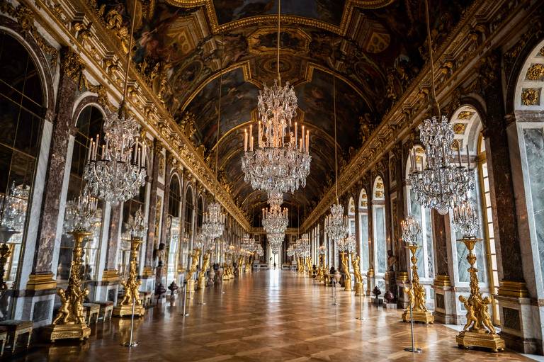 A imagem mostra o Salão de Espelhos do Palácio de Versalhes, com um longo corredor adornado por lustres de cristal e colunas douradas. As paredes são decoradas com espelhos e detalhes em mármore, enquanto o teto apresenta pinturas elaboradas. O chão é de madeira com um padrão geométrico.
