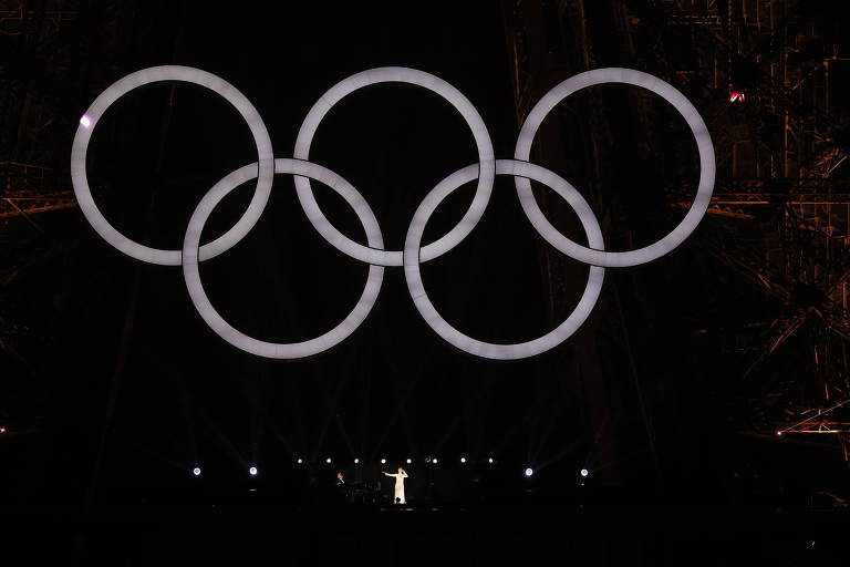 A imagem mostra os cinco anéis olímpicos, iluminados em um fundo escuro. Abaixo dos anéis, há uma figura iluminada, em um ambiente que parece ser um evento ou cerimônia relacionada aos Jogos Olímpicos.