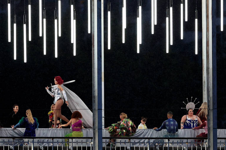 A imagem mostra um desfile de moda noturno com várias pessoas em um palco. Uma modelo se destaca à frente, vestindo um traje branco e uma capa, enquanto outras pessoas estão sentadas ao fundo, algumas com roupas coloridas e elaboradas. O ambiente é iluminado por lâmpadas pendentes que criam um efeito dramático no cenário escuro.
