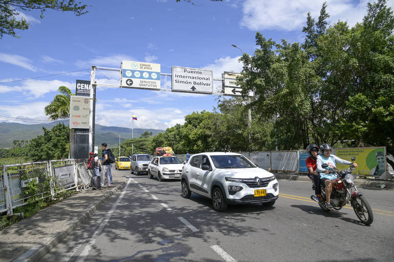 A imagem mostra um cruzamento de estrada com veículos em movimento, incluindo carros e uma motocicleta. Há pessoas caminhando ao lado da estrada e placas de sinalização visíveis ao fundo. O céu está claro com algumas nuvens e há vegetação ao redor.