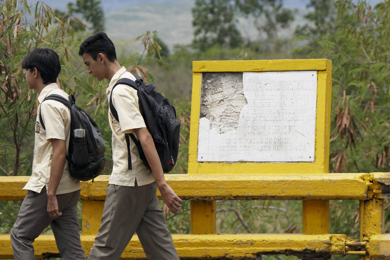 Dois estudantes em uniformes escolares caminham em uma estrada. Eles estão usando mochilas e passam ao lado de um painel amarelo, que contém um documento ou aviso, mas está parcialmente danificado. Ao fundo, há vegetação e uma paisagem montanhosa.