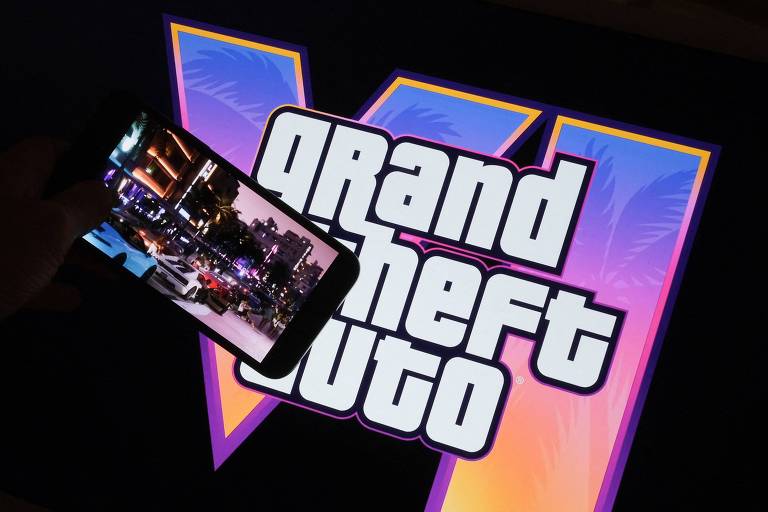 A imagem mostra um smartphone posicionado sobre uma superfície que exibe o logotipo do jogo 'Grand Theft Auto VI'. O logotipo é grande e colorido, com um fundo em tons de azul e rosa. O smartphone exibe uma imagem de um cenário urbano, possivelmente do próprio jogo.
