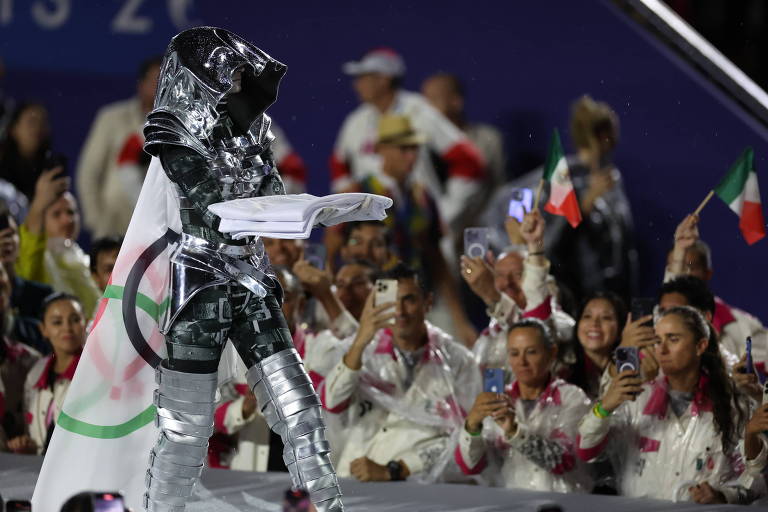 A imagem mostra um astronauta vestido com um traje prateado e um capacete, segurando um objeto em suas mãos. Ao fundo, um grupo de pessoas vestidas com roupas brancas e detalhes coloridos, segurando bandeiras, observa e aplaude. A atmosfera é de celebração e entusiasmo.