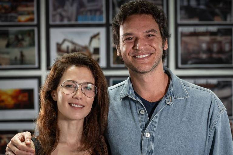 A imagem mostra duas pessoas sorrindo em um estúdio. A mulher à esquerda tem cabelo castanho claro e usa óculos, vestindo uma camiseta preta com a inscrição 'Les Femmes'. O homem à direita tem cabelo escuro e usa uma camisa de jeans. Ao fundo, há várias fotografias emolduradas nas paredes.