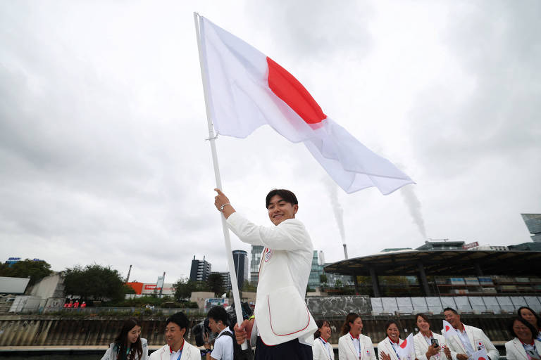 Um homem está segurando uma bandeira do Japão, que é branca com um círculo vermelho no centro. Ele está sorrindo e vestido com um traje claro. Ao fundo, há um grupo de pessoas vestindo roupas brancas, e o céu está nublado.