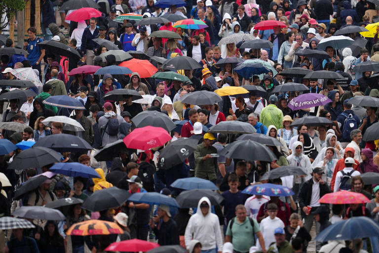 A imagem mostra uma grande multidão de pessoas caminhando sob a chuva, todas segurando guarda-chuvas de diversas cores. A cena é cheia de movimento, com pessoas vestindo roupas de diferentes estilos, algumas com capuzes e outras com chapéus. O ambiente parece ser urbano, e a atmosfera é de um dia chuvoso.
