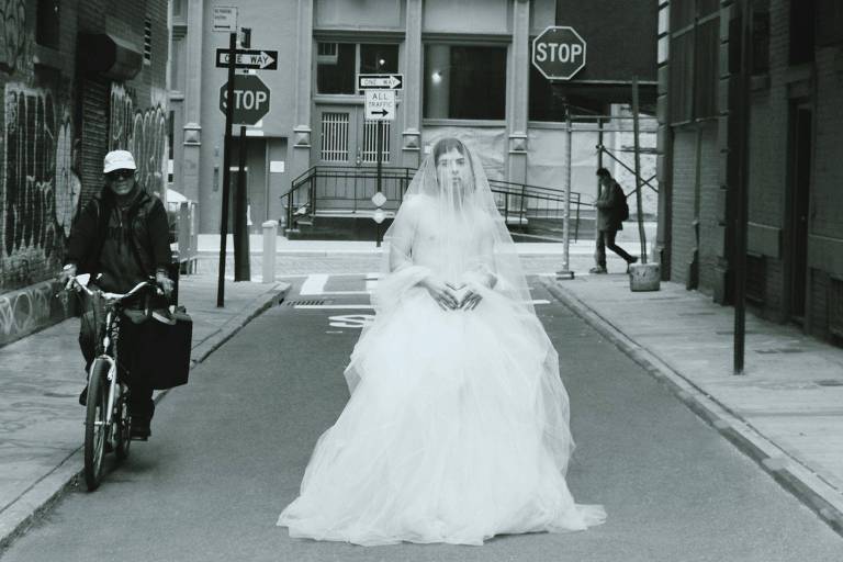 Na foto em preto e branco, uma pessoa está no meio de uma ra usando um vestido de noiva. Do seu lado, passa uma pessoa de bicicleta. Ao fundo, duas placas de Pare e uma pessoa atravessando uma esquina