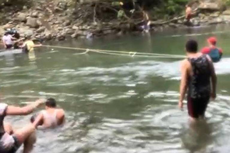 Na foto, uma corda está estendida sobre um rio. Pessoas próximas a ela usam a corda para facilitar a caminhada de uma margem para outra
