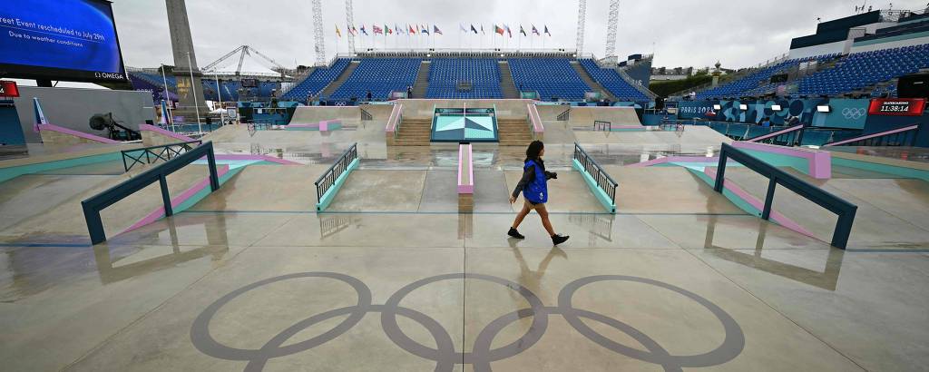 A imagem mostra uma área das instalações olímpicas com um piso molhado refletindo os cinco anéis olímpicos. Uma pessoa caminha pelo local, que parece estar sob um céu nublado. Ao fundo, há estruturas e telões visíveis, indicando que é um espaço preparado para eventos esportivos.