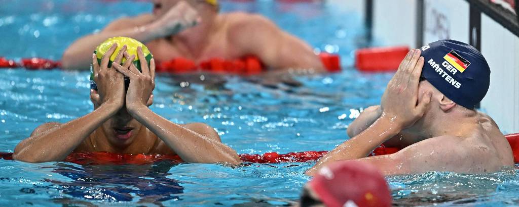 A imagem mostra nadadores em uma piscina, alguns com toucas de natação. Dois nadadores estão com as mãos no rosto, enquanto outros estão ao fundo. A água da piscina é azul e há boias vermelhas ao redor das raias.