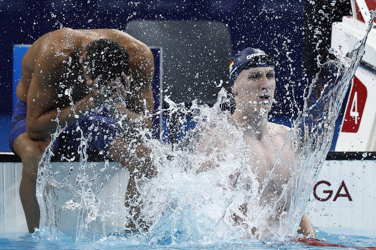 A imagem mostra dois nadadores em uma piscina. Um nadador está sentado na borda, com uma perna pendurada na água, enquanto o outro está parcialmente submerso, com água espirrando ao seu redor. O fundo é azul, refletindo a água da piscina.
