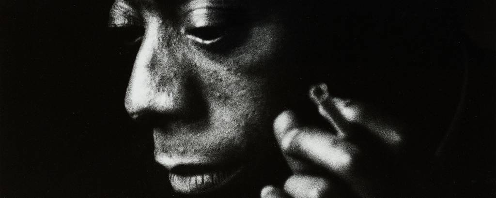 foto em preto e branco, sombreada, de homem negro olhando para baixo e para a esquerda, com um cigarro entre os dedos