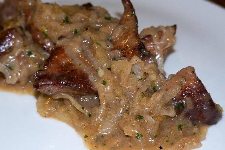 A imagem mostra um prato com carne cozida, coberta por um molho espesso e cremoso. A carne apresenta uma coloração marrom escura, e o molho tem uma tonalidade mais clara, com pedaços visíveis de temperos e ervas.