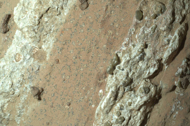 Imagem mostrando a superfície de Marte, com uma textura irregular e rochas visíveis. A cor predominante é um tom marrom claro, com áreas mais claras e algumas pedras esparsas.