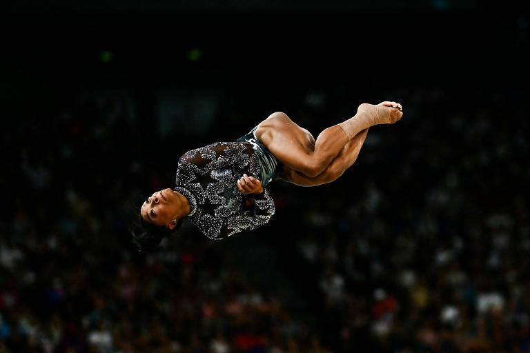 Uma ginasta está realizando um salto acrobático no ar, com o corpo em posição invertida. Ela usa um traje de competição escuro e está cercada por uma multidão que assiste ao evento. A imagem captura o momento em que ela está prestes a aterrissar.
