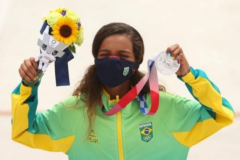 Rayssa Leal segurando sua medalha de prata nos Jogos Olimpicos de Tóquio 2021, ela usa máscara contra a covid-19, e segura na outra mão um pequeno buquê de flores, onde se destaca um girassol