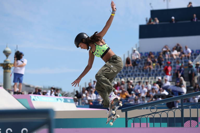 Uma skatista está realizando uma manobra em uma pista de skate. Ela usa uma blusa verde e calças camufladas, enquanto salta sobre uma borda. Ao fundo, há uma plateia assistindo ao evento, com várias pessoas visíveis em um ambiente ao ar livre sob um céu claro.