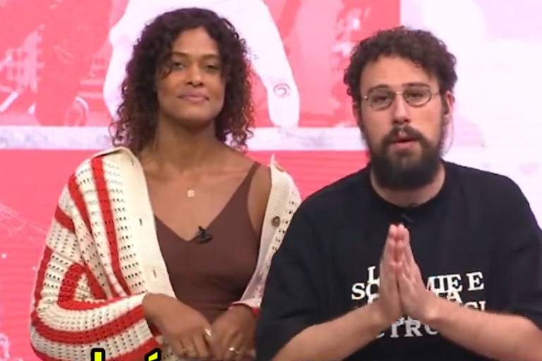 A imagem mostra dois apresentadores em um estúdio. À esquerda, uma mulher com cabelo cacheado e solto, vestindo uma blusa marrom e um cardigan branco com listras vermelhas. À direita, um homem com barba e óculos, usando uma camiseta preta com texto em branco. O fundo é uma tela com cores vermelhas e imagens desfocadas.