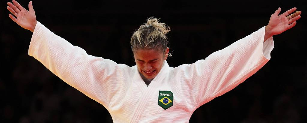 Uma atleta de judô com os braços levantados e a cabeça inclinada para baixo. Ela veste um judogi branco com uma faixa preta e um emblema verde no peito. O fundo é desfocado, sugerindo um ambiente de competição.
