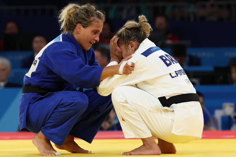 Duas atletas de judô estão em uma competição. A atleta à esquerda veste um judogi azul e está consolando a atleta à direita, vestindo um judogi branco, com a cabeça baixa e as mãos na frente do rosto. O fundo mostra uma plateia assistindo ao evento.
