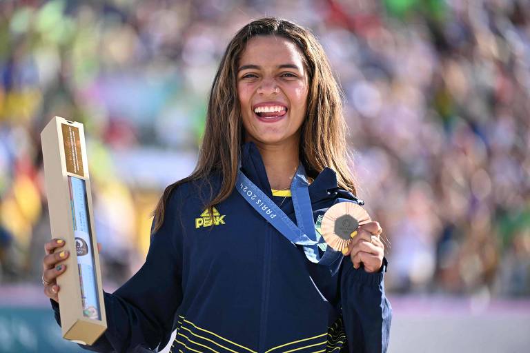 Uma atleta brasileira está em um pódio, segurando um troféu e um boneco. Ela usa uma capa com as cores da bandeira do Brasil e exibe uma medalha de bronze ao redor do pescoço. O fundo mostra uma multidão e uma estrutura de evento esportivo.
