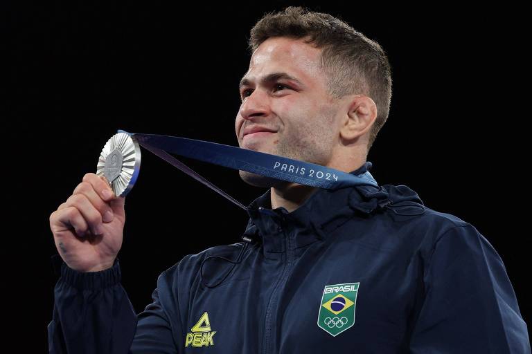 Um atleta está sorrindo enquanto segura uma medalha de prata com uma fita azul. Ele está usando um casaco escuro com o emblema do Brasil. O fundo é escuro, destacando a expressão de felicidade do atleta.

