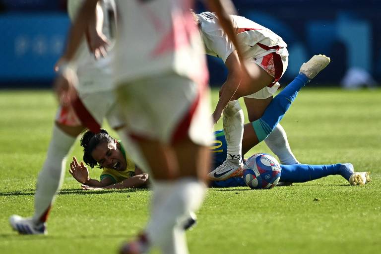 A imagem mostra uma cena de um jogo de futebol, onde uma jogadora está caída no gramado, enquanto outras jogadoras estão em pé, alguns deles sobre o caído. O jogador no chão parece estar em uma posição vulnerável, e a ação sugere um momento intenso do jogo.