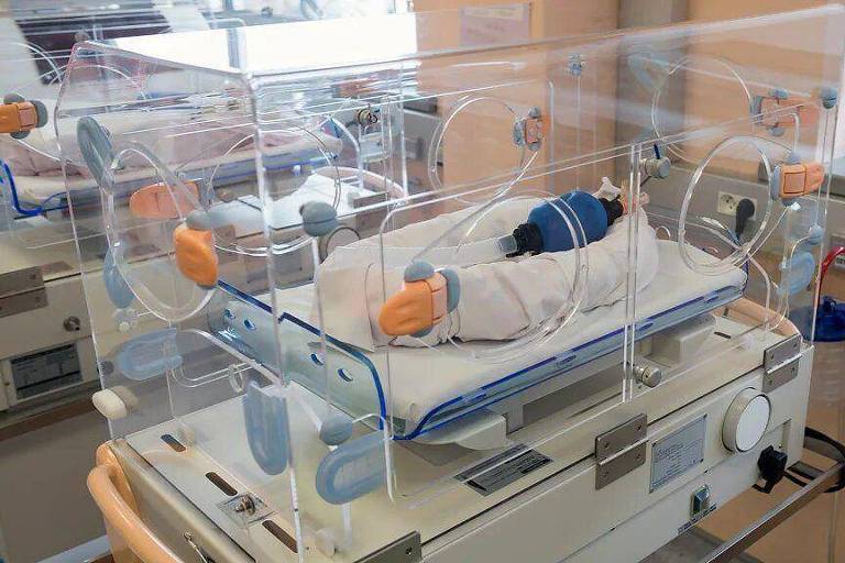 A imagem mostra uma incubadora em uma unidade de terapia intensiva neonatal. A incubadora é transparente e possui aberturas vedadas por onde passam tubos e fios médicos.