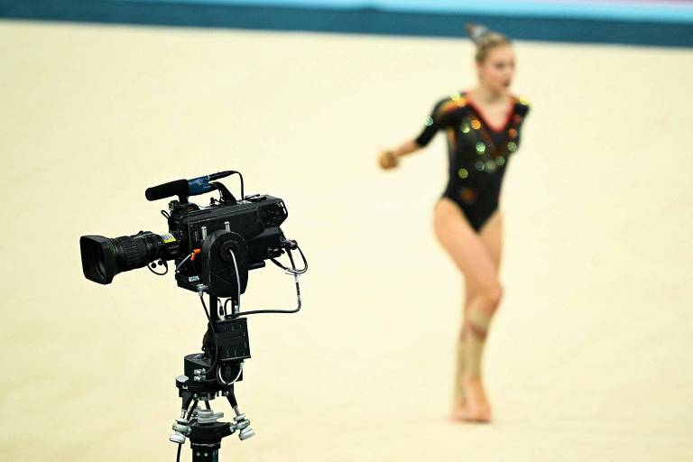 A imagem mostra uma câmera de vídeo em primeiro plano, posicionada em um tripé, filmando uma ginasta em movimento ao fundo. A ginasta está vestindo um traje de competição preto com detalhes brilhantes e está em uma pose dinâmica. O fundo é uma superfície de ginástica clara.
