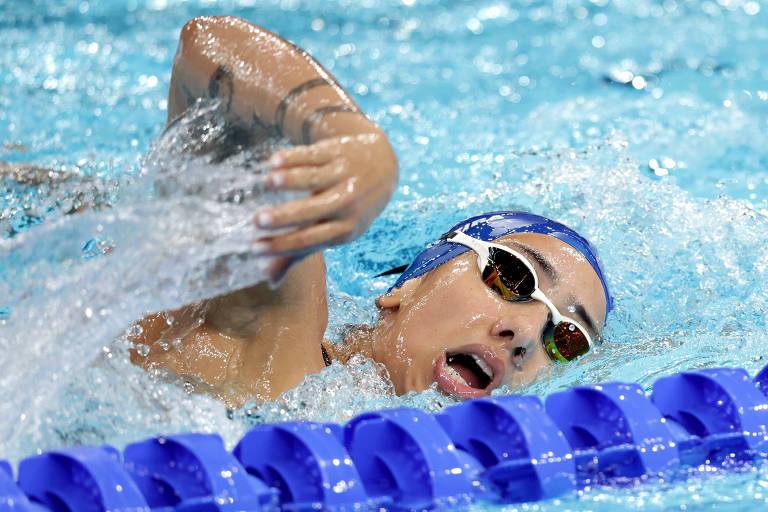 Uma nadadora está competindo em uma piscina, realizando um estilo livre. Ela usa um touca azul e óculos de natação. A água está espirrando ao redor dela enquanto ela se move rapidamente, e a borda da piscina é visível com boias azuis.
