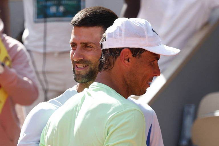 A imagem mostra dois jogadores de tênis se cumprimentando após uma partida. Um deles está usando uma camiseta branca e o outro uma camiseta verde claro, com um boné branco. Ambos estão sorrindo e parecem estar em um momento amigável. Ao fundo, é possível ver espectadores e outros jogadores.