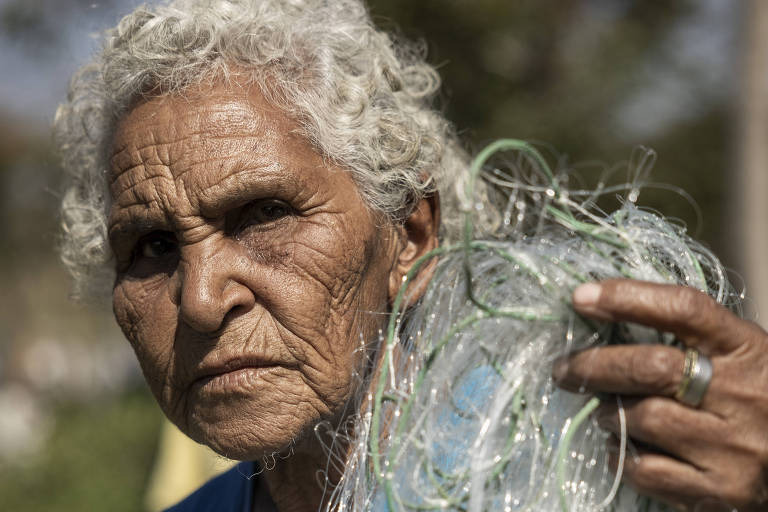 Sebastiana é uma mulher idosa, pela enrugada e parda. Tem pelos 70 anos, os gabelos grisalhos. Segura uma rede de pesca com a mão esquerda enquanto olha para a câmera. Veste uma camiseta azul claro