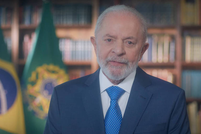 Homem branco, com cabelo e barba brancas, veste terno e gravata. ao fundo a bandeira do Brasil e estante com livros
