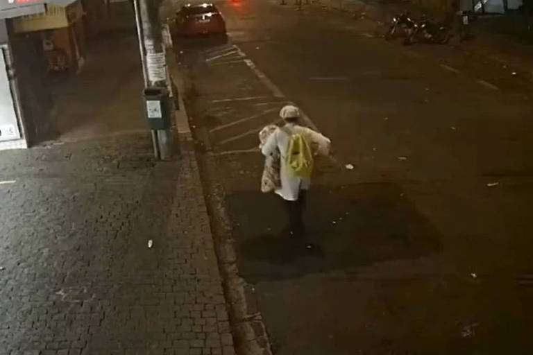 A imagem mostra uma pessoa caminhando em uma rua à noite. A pessoa está vestindo uma camisa clara e um boné, e carrega uma mochila amarela. O ambiente é urbano, com algumas luzes ao fundo e veículos estacionados. O chão é pavimentado e há lixo espalhado na calçada.