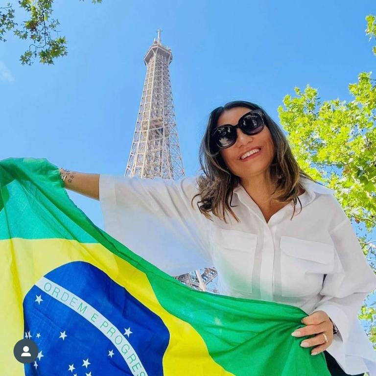 A imagem mostra uma mulher sorridente segurando a bandeira do Brasil em frente à Torre Eiffel. Ela está vestindo uma camisa branca e óculos escuros. O céu está claro e azul, com algumas folhas verdes visíveis ao redor.