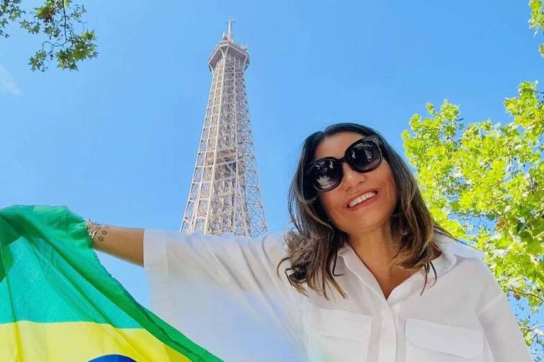 A imagem mostra uma mulher sorridente segurando a bandeira do Brasil em frente à Torre Eiffel. Ela está vestindo uma camisa branca e óculos escuros. O céu está claro e azul, com algumas folhas verdes visíveis ao redor.