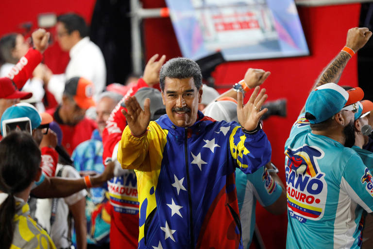 A imagem mostra um grupo de pessoas em um evento, com destaque para um homem no centro que está usando uma camisa com as cores da bandeira da Venezuela. Ele está sorrindo e acenando, enquanto outras pessoas ao redor também estão animadas, levantando os braços. O ambiente parece festivo, com bandeiras e roupas coloridas.